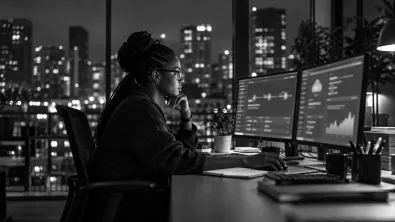 An engineer working late at two monitors, with city lights through the window behind her