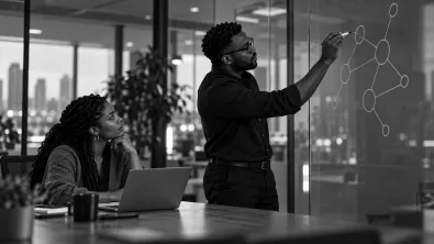 An engineer drawing a diagram of connected circles on a glass wall while a colleague at a laptop looks on